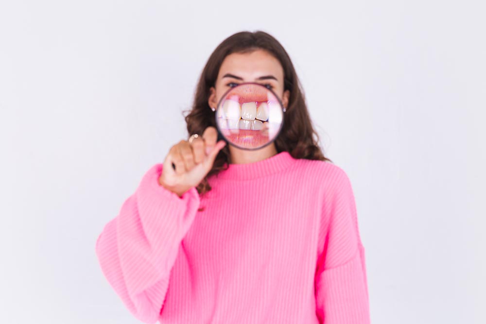 young beautiful woman with freckles light makeup sweater white wall with magnifier shows white teeth perfect smile